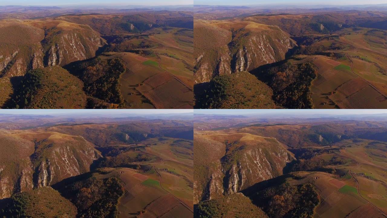 在阳光明媚的秋天，在散布着房屋和橙色落叶松森林的小山村上空前进。山间蜿蜒的山路鸟瞰图。