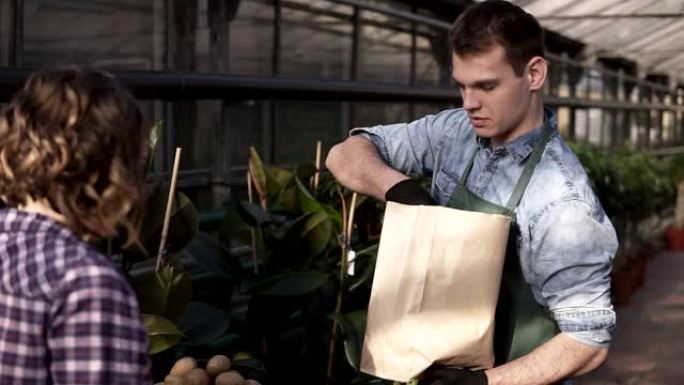 高加索人，穿着绿色围裙的高个子推销员正在将新鲜的沙拉，胡萝卜放在纸袋中给温室市场的顾客。微笑的家伙卖