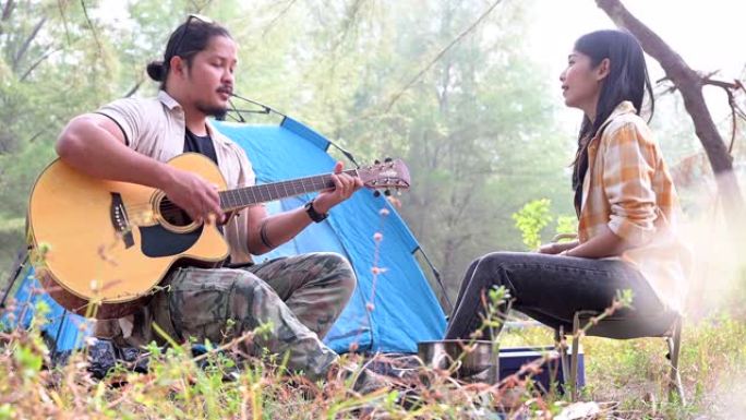 亚洲年轻夫妇喜欢在松林露营，喜欢运动和情侣关系，弹吉他，夏天在露营帐篷前玩得开心。