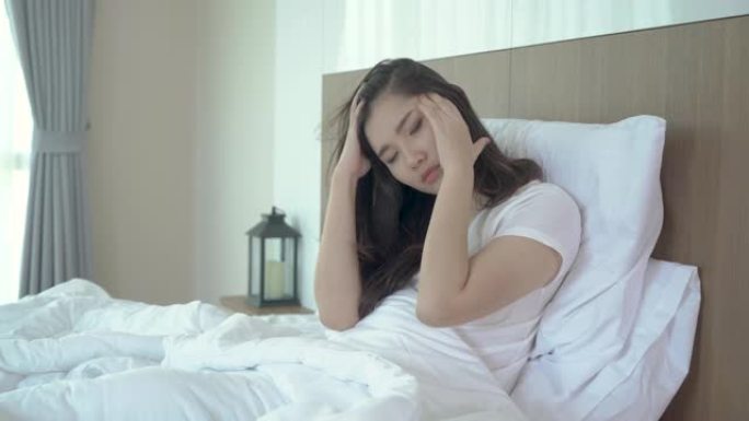 迷人的年轻亚洲妇女醒来在她的床上，握着他们的手从头痛，看起来不开心，感觉头痛/偏头痛/压力/生病。妇