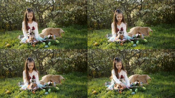 院子里的女孩坐在草地上开花树木的背景上，用彩蛋装饰复活节兔子。