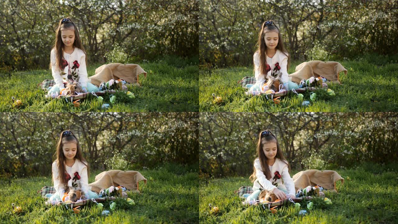 院子里的女孩坐在草地上开花树木的背景上，用彩蛋装饰复活节兔子。