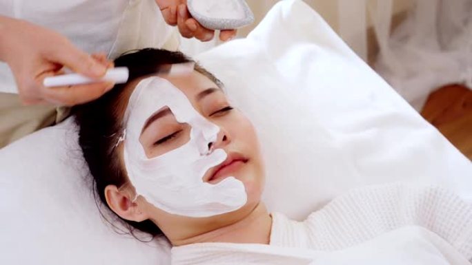 美容中心，亚洲美容治疗妇女舒适放松，而美容师的手使用刷子在脸上使用带有水疗霜的口罩来护理脸部