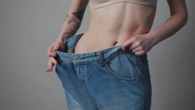 减肥和身体形象概念。年轻女子显示她的体重减轻，穿着她的旧牛仔裤。穿着大牛仔裤的苗条女孩展示了她开始吃