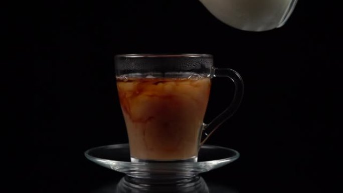 在玻璃茶杯和茶托中的红茶中，从黑色背景上的玻璃壶中倒牛奶，慢动作拍摄