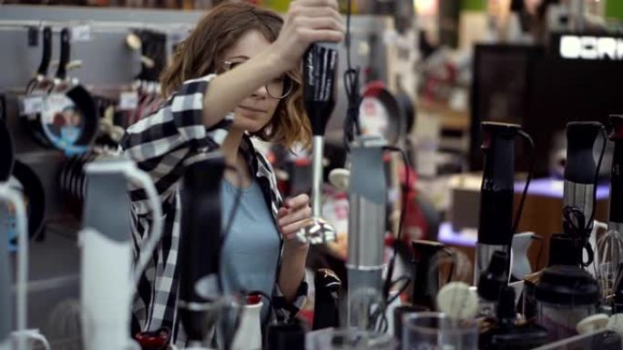 在电器商店中，一位穿着格子衬衫的迷人卷发女人通过查看并握住手中的设备来选择搅拌器棒。慢动作
