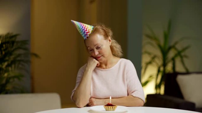 戴党帽的悲伤的高级女人独自坐在公寓的桌子上，手放在下巴上，看着蜡烛燃烧的生日蛋糕，感到孤独和沮丧