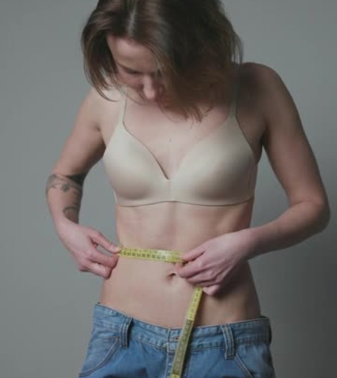穿着牛仔裤站在镜子前的女人减肥后正在测量腰围。减肥和节食概念。垂直射击