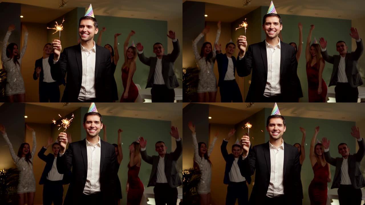 视频肖像，快乐的年轻人戴着派对帽子，拿着烟火，看着相机，微笑着，而他的朋友们在家里快乐的庆祝派对上跳