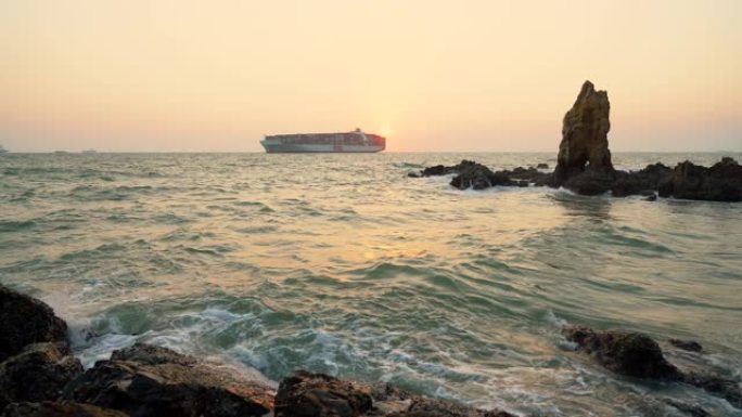 日落时与货船一起淹没在海中的大型岩石。海浪溅到淹没的岩石上。