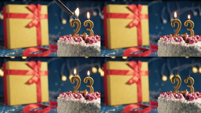 白色生日蛋糕编号23点灯燃烧的金色蜡烛，带灯的蓝色背景和用红丝带绑起来的礼物黄色盒子。特写
