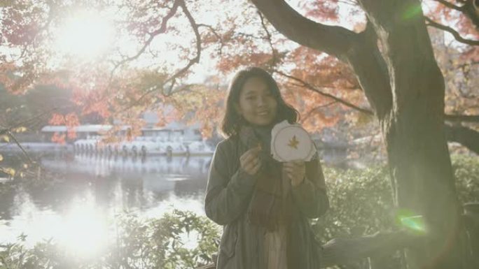 在阳光明媚的日子里，在东京市的公园里，微笑的年轻亚洲妇女拿着带有枫叶图案的刺绣框的肖像。