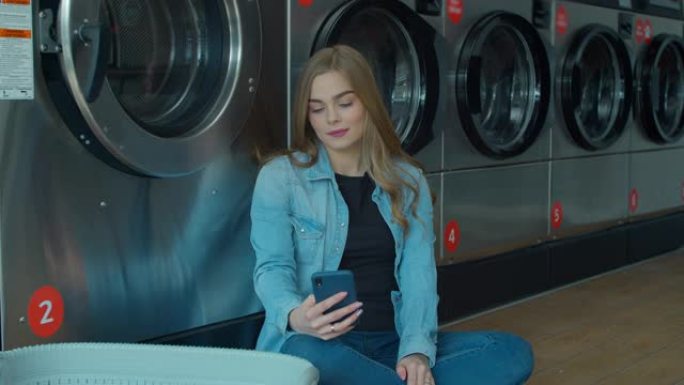 等待洗衣服的年轻女子坐在地板上，在自助洗衣店使用智能手机。
