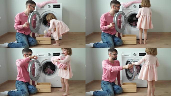 快乐的年轻父亲带着微笑的3岁女儿把毛绒玩具放在洗衣机里洗。女孩关闭舱门，打开家用电器。