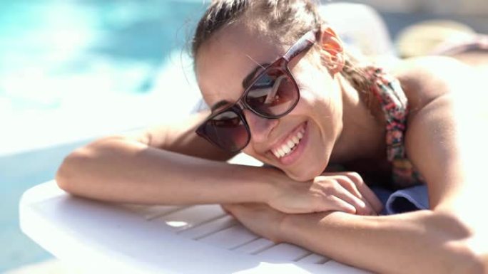 近距离的美丽微笑的白人女孩戴着太阳镜躺在泳池边的桌子上晒太阳