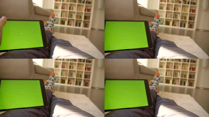 男性腿的特写镜头悠闲地坐在地毯上的地板上，双手拿着平板电脑，背景是绿色色度屏幕，背景是书架