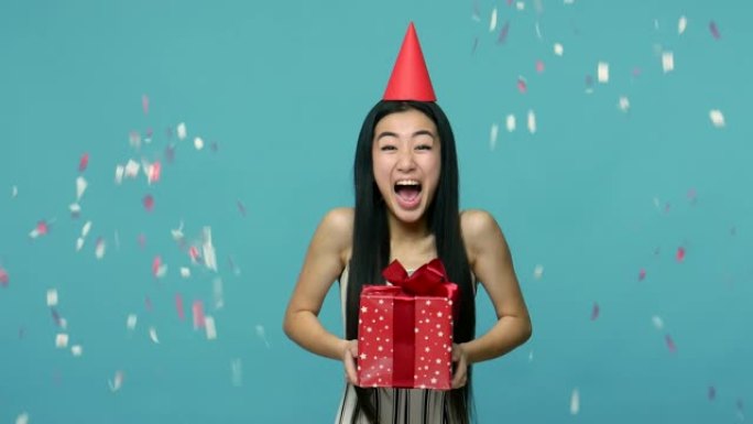 五彩纸屑落在兴奋的亚洲女人身上，头上有滑稽的派对锥，女孩拿着礼物盒子，接受问候