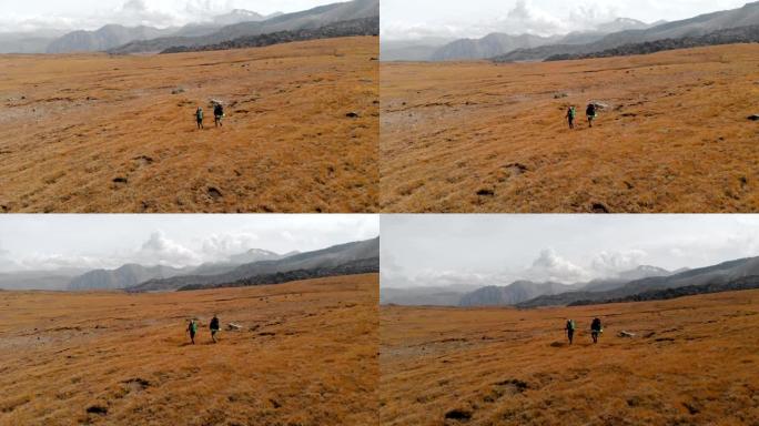 一对带着帽子和墨镜的大背包的旅行者男人和女人的鸟瞰图沿着被史诗般的山脉环绕的高山高原行走