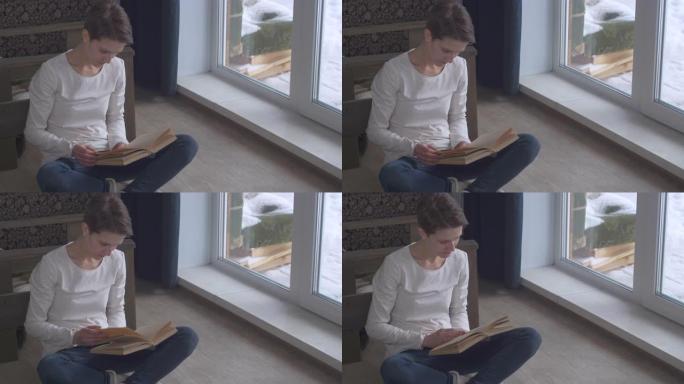 在一个寒冷的冬日傍晚，一个短发的年轻女孩坐在窗边的地板上看书。享受文学的女人。女性教育和学习。小屋的
