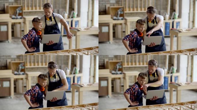 中年木匠和他的小儿子在木工店里一起建造木船模型。父亲在笔记本电脑上向孩子展示计划并解释细节
