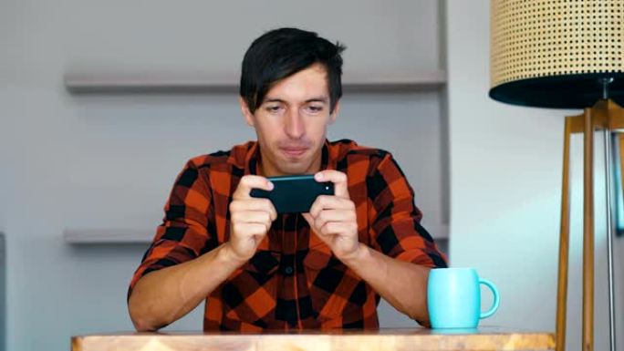 拿着智能手机玩手机游戏的年轻人坐在家里的桌子上，快乐的年轻人使用社交媒体应用程序在手机上工作学习看着