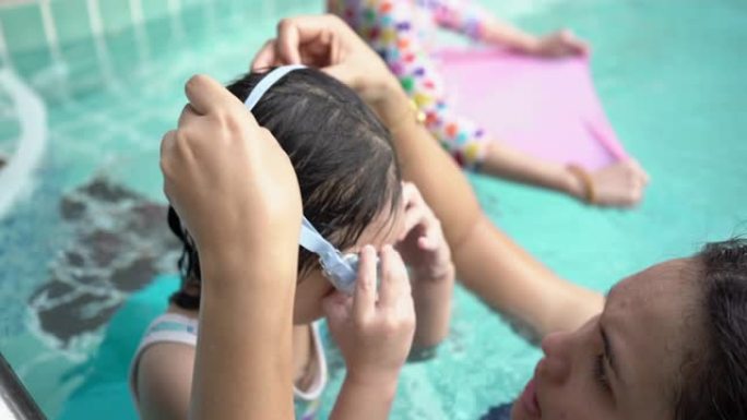 母亲为女儿戴了泳镜。亚洲年轻的母亲和可爱的卷发小女孩在游泳池里玩得开心，幸福的家庭理念。手持镜头，真