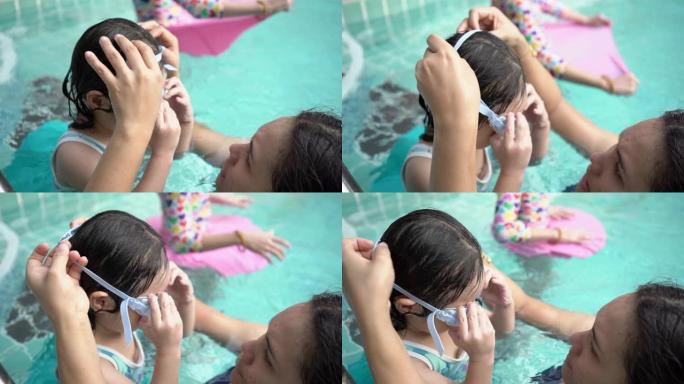 母亲为女儿戴了泳镜。亚洲年轻的母亲和可爱的卷发小女孩在游泳池里玩得开心，幸福的家庭理念。手持镜头，真