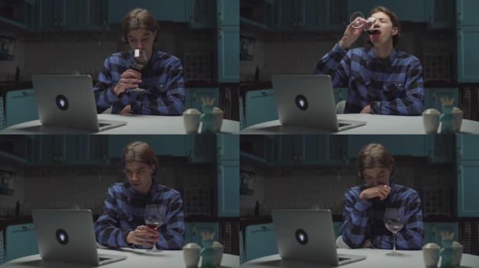绝望的年轻人喝红酒，看着在线笔记本电脑独自坐在厨房里。悲伤的男性在家喝酒。