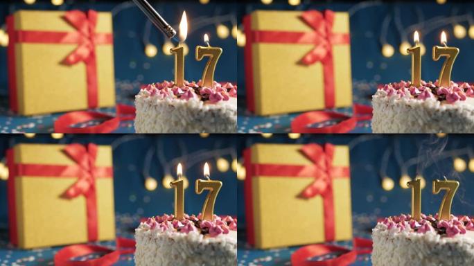 白色生日蛋糕编号17点灯燃烧的金色蜡烛，带灯的蓝色背景和用红丝带绑起来的礼物黄色盒子。特写