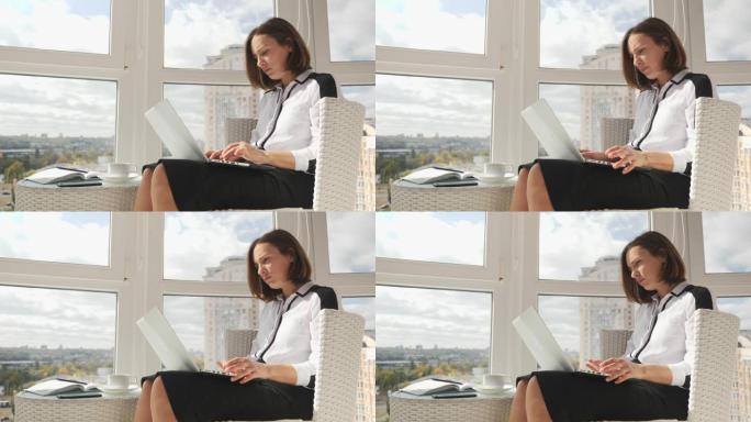 穿着白色衬衫和黑色裙子的聪明成功的年轻女商人在大窗户的工作场所在笔记本电脑上工作