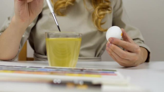 面目全非的女人用刷子画复活节彩蛋。桌子上是一杯水和一碗鸡蛋。为假期做准备