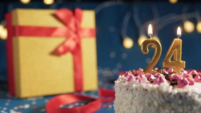 白色生日蛋糕编号24点灯燃烧的金色蜡烛，带灯的蓝色背景和用红丝带绑起来的礼物黄色盒子。特写