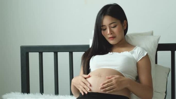 亚洲美丽的年轻女子在白色床上抚摸她的腹部，小心翼翼地爱着子宫里的婴儿