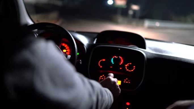 无法识别的驾驶员在夜间城市街道上握着方向盘并驾驶汽车。从汽车驾驶室观看霓虹灯照亮汽车仪表板，从车窗眩