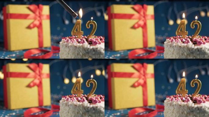 白色生日蛋糕编号42点灯燃烧的金色蜡烛，蓝色背景带灯和用红丝带绑起来的礼物黄色盒子。特写