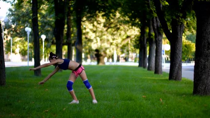 在阳光明媚的夏日，在城市公园的草地上，穿着运动服的七年美丽的白人女孩在草地上练习车轮体操运动。慢动作