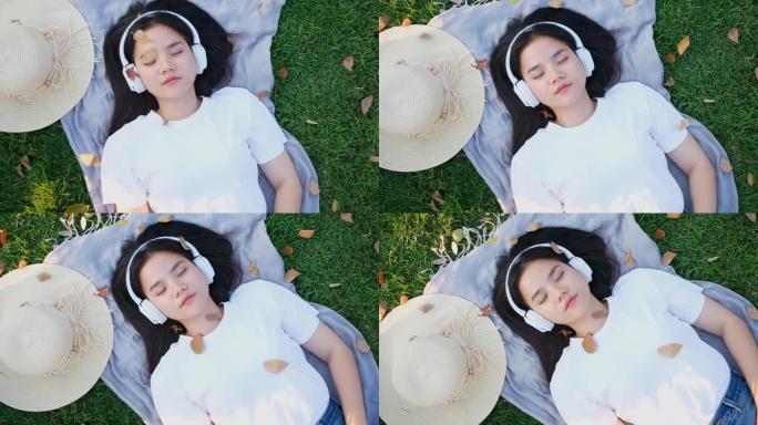 亚洲女人通过耳机听音乐睡觉时的慢动作，亚洲女孩夏天戴着耳机在公园地上打盹躺着，现代科技和人们的生活方