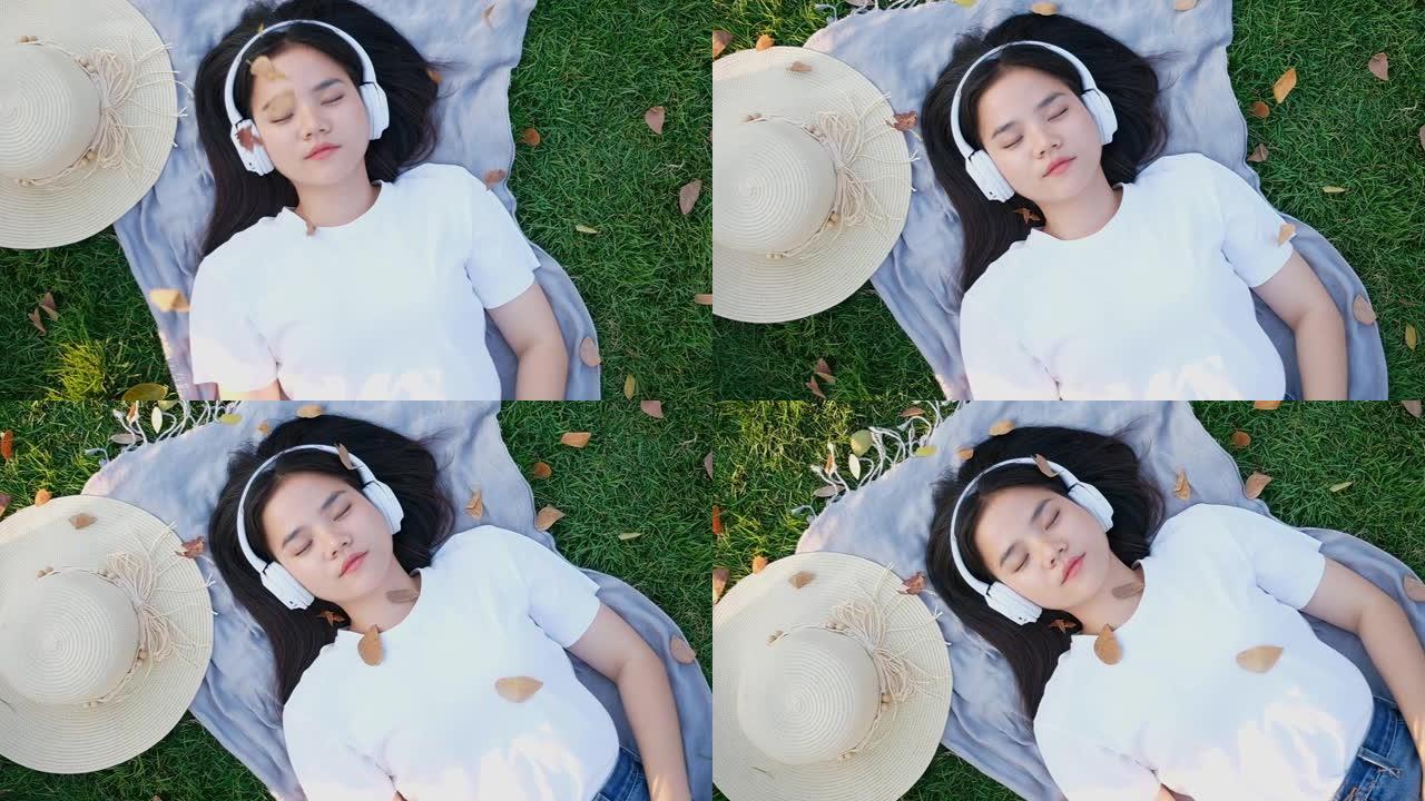 亚洲女人通过耳机听音乐睡觉时的慢动作，亚洲女孩夏天戴着耳机在公园地上打盹躺着，现代科技和人们的生活方