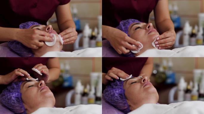 美容师用海绵清洁白人妇女的脸。闭着眼睛躺在沙发上的病人