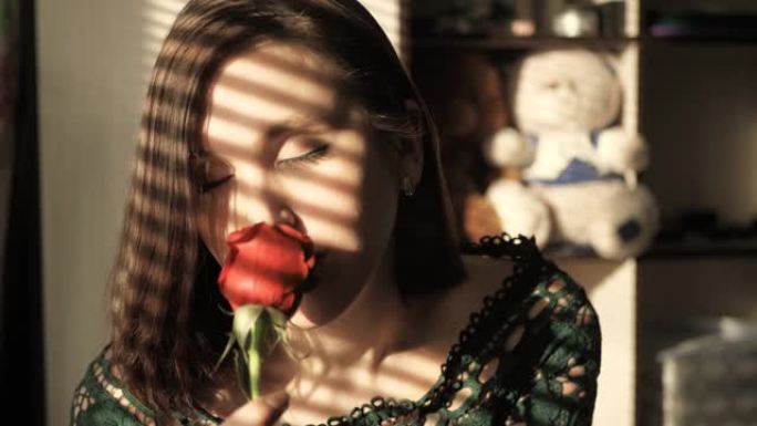 美丽的女孩坐在日落的窗边，爱着红玫瑰，嗅着花。情人节，2月14日，生日，周年纪念日，第一次约会的概念