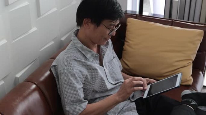 资深亚洲男子在家里的客厅沙发上玩平板电脑，亚洲老人在平板电脑上通过视频通话放松和幸福