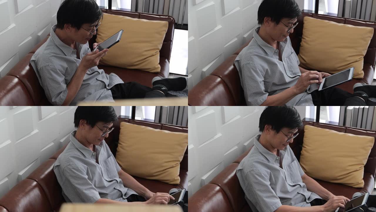 资深亚洲男子在家里的客厅沙发上玩平板电脑，亚洲老人在平板电脑上通过视频通话放松和幸福