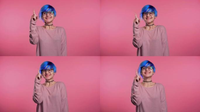 年轻思考思考的女人的肖像，有着不同寻常的蓝色发型，有想法的瞬间指向粉红色工作室背景。微笑快乐的女孩表