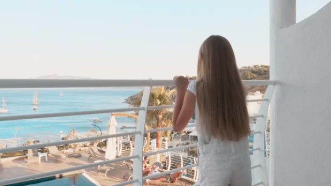 度假时，旅游女孩从度假酒店的房间出来到阳台。少女从酒店露台观看海景。游泳池背景阳台上的年轻女孩。