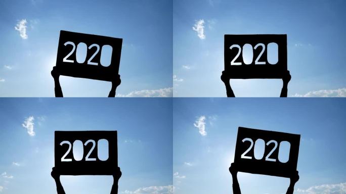 手握回收纸板的剪影，2020文字在蓝天背景和阳光照耀着文字。新年和庆祝活动的概念。