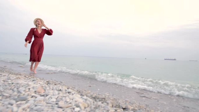 日落时分，一个穿着红色浅色连衣裙和草帽的年轻迷人的微笑女孩沿着海边散步。轻盈与美的女性气质与独立性