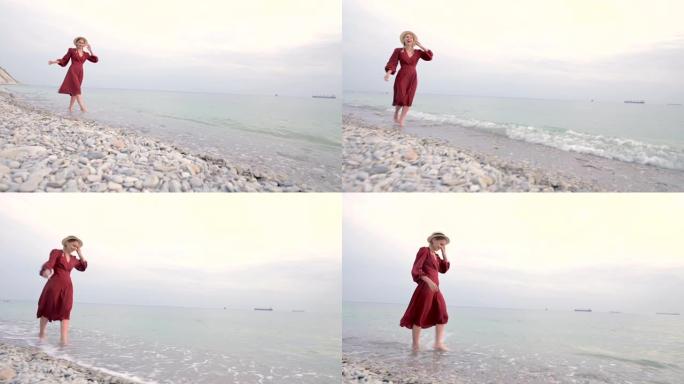 日落时分，一个穿着红色浅色连衣裙和草帽的年轻迷人的微笑女孩沿着海边散步。轻盈与美的女性气质与独立性