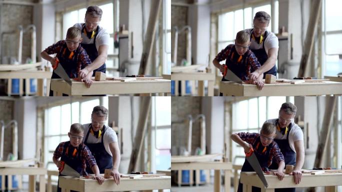 中年木匠的中等镜头教小儿子如何使用手锯切割站在木工车间工作台上的木板，穿着围裙和防护眼镜