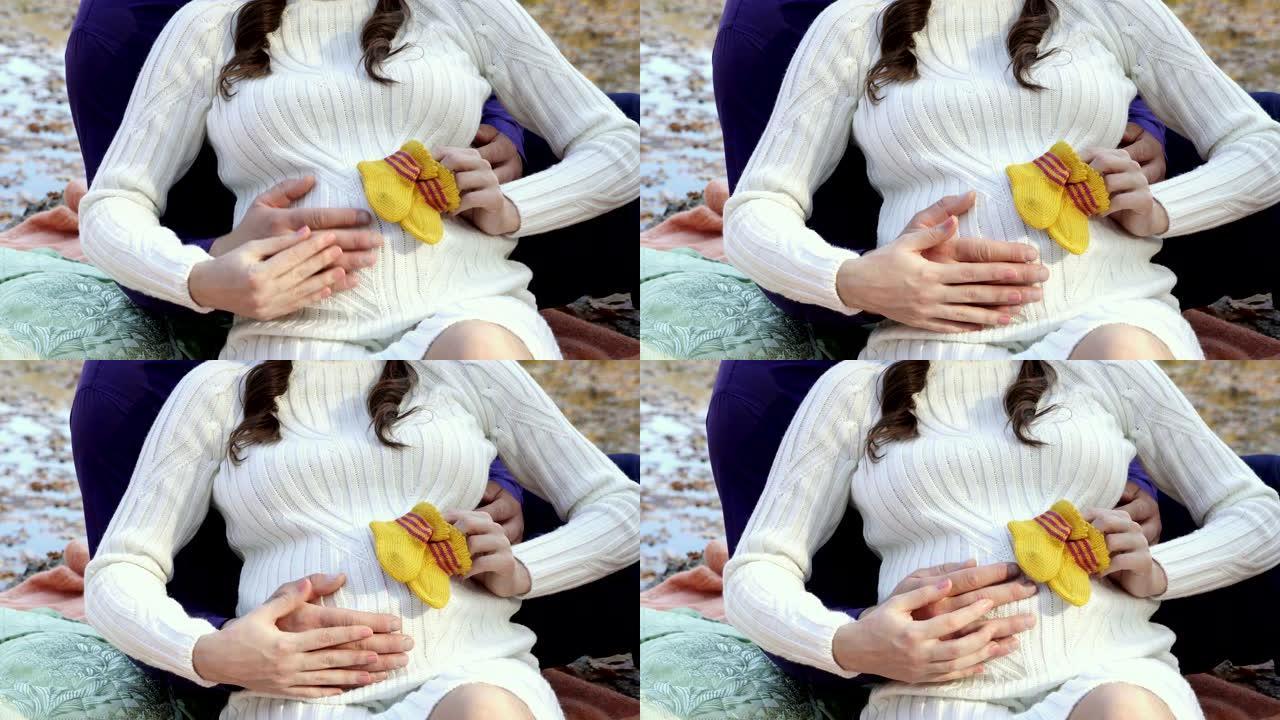 特写镜头，一只男女之手抚摸着一名孕妇的腹部，该孕妇在阳光明媚的日子里穿着白色针织毛衣坐在大自然中，并