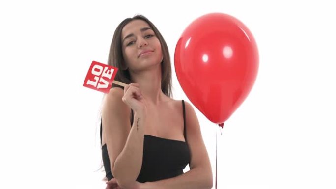 年轻漂亮的女人拿着爱字的木板，对着镜头摆姿势。模型站在充满氦气的红色气球附近。情人节概念